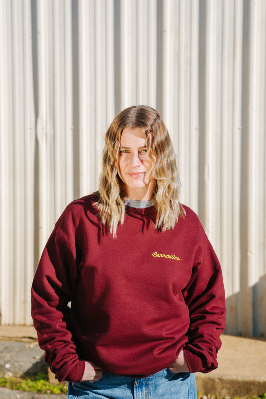 A person wearing a maroon crewneck sweatshirt with 'Bonnevilles' embroidered on the top left, standing in front of a corrugated metal wall.