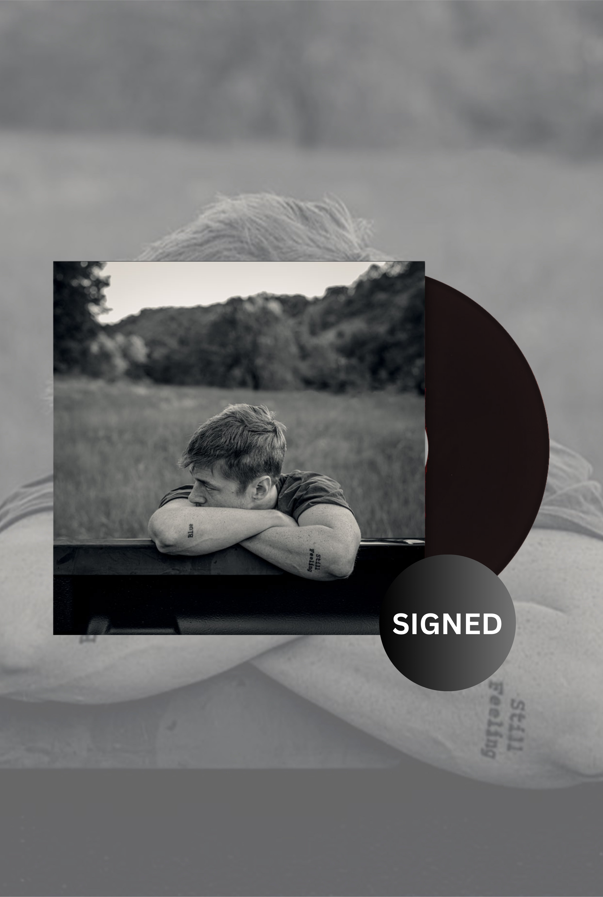 A black and white image of a vinyl LP with a 'Signed' label, showing a person lying down next to a record.